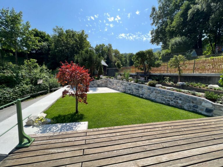 Ein gepflegter Garten mit einem roten Baum, Steinmauern und einer Holzterrasse bei Tageslicht